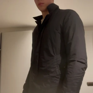 Svart jacka från Aspesi - Säljer en stilren svart jacka från Aspesi med orange detaljer och diskret logga på axeln. Jackan har klassisk krage, raka linjer och knäppning framtill. Tillverkad i lätt polyester, perfekt för höst och vår. Snygg och enkel att matcha med det mesta.