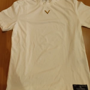Vit t-shirt från Valentino med V-detalj - Snygg vit t-shirt från Valentino, tillverkad i Italien. T-shirten har en stilren V-logga i silver på bröstet samt en klassisk rund hals. Materialet är mjuk bomull och passformen är normal. Perfekt för dig som gillar exklusiva märken och minimalistisk design. Helt ny fast utan prislapp. Köptes i Rom. Tveka inte att höra av dig om du har frågor och priset kan diskuteras.