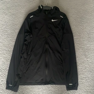 Svart Nike miler  från Nike med huva  - Aldrig använt. Helt ny. Svart Nike miler från Nike med hel dragkedja, huva och reflekterande detaljer. Jackan har lång ärm och är tillverkad i lätt polyester, perfekt för träning eller när du vill ha en sportig look. Swoosh-logga på bröstet och diskreta detaljer på axlar och ärmslut.