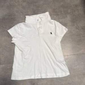 Vit pikétröja från Ralph Lauren - Vit pikétröja från Ralph Lauren med klassisk krage och korta ärmar. Tröjan har knappar framtill och den ikoniska broderade loggan i svart på bröstet. Tillverkad i mjuk bomull som är skön mot huden. Perfekt för en clean och stilren look.