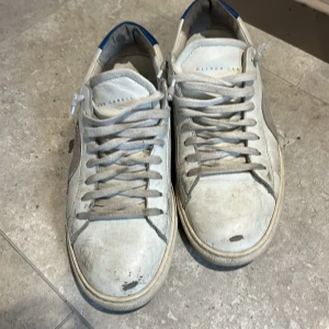 Oliver Cabell vita sneakers med blå detalj - Säljer ett par vita sneakers från Oliver Cabell med grå cirkeldetalj på sidan och blå häl. Skorna har snörning, platt sula och rund tå. Tillverkade i skinn med coola röda stygn på sidan. Perfekt för dig som gillar stilrena och unika sneakers.