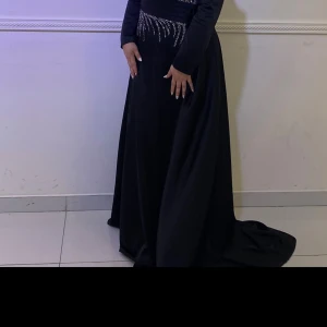 Svart långklänning med glitterdetaljer - Svart långklänning med lång ärm och glittriga silverdetaljer över midjan och bröstet. Klänningen har en elegant, svepande form och är tillverkad i ett slätt material som ger ett lyxigt intryck. Perfekt för bal eller fest, med snygga veck vid midjan och diskret glans.