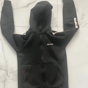 Svart hoodie från BOSS med huva - Svart hoodie från BOSS med dragkedja, huva och två fickor med zip framtill. Snygga beige och silverfärgade ränder längs ärmarna ger en sportig vibe. BOSS-logga på bröstet. Perfekt för chill eller träning.