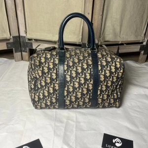 Dior Boston - Snygg handväska från Dior med klassiskt blått monogrammönster på beige canvas och mörkblå skinnhandtag. Väskan har rundad form, dragkedja upptill och detaljer i läder. Perfekt för dig som vill ha en ikonisk och trendig accessoar.