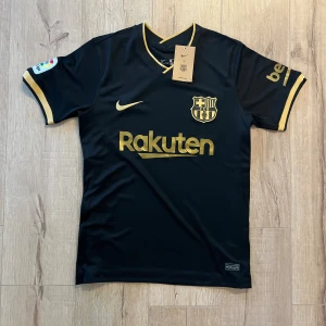 Svart FC Barcelona fotbollströja Nike - Snygg svart FC Barcelona matchtröja från Nike med guldiga detaljer och tryck. Kortärmad med V-ringad hals och La Liga-märke på ärmen. Materialet är lätt och andas, perfekt för träning eller att bära på läktaren. Rakuten och UNICEF-loggor i guld framtill och baktill.