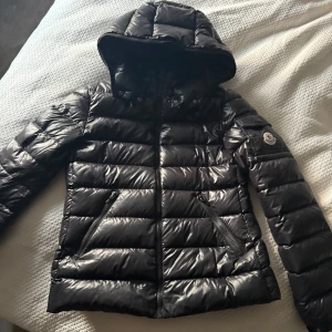 Äkta moncler jacka  - Säljer en svart, glansig dunjacka från Moncler med luva och dragkedjor på fickorna. Jackan har klassisk design, lång ärm och ikonisk Moncler- logga på ärmen. Perfekt för kalla dagar. Köpt i monclers egna butik. Modell Bady Hooded Short Down Jacket. Den går in lite i midjan och formar sig efter kroppen vilket är jätte snyggt. Den har tyvärr blivit för liten på mig, den har ett litet typ rivmärke vid armen som knappt syns, se bild 5. Skriv för flera bilder. Stolek 14