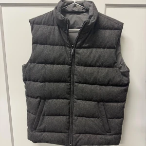 Mörkgrå dunväst från Volt - Snygg mörkgrå dunväst från Volt. Ena sidan har en mjuk ull-liknande yta och den andra är slätare i syntetmaterial, vilket gör västen vändbar. Perfekt för lager-på-lager under kyliga dagar.