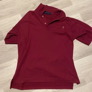 Vinröd pikétröja från Ralph Lauren - Snygg vinröd pikétröja från Ralph Lauren med klassisk krage och två knappar framtill. Ikoniska broderade loggan på bröstet. Perfekt basic med korta ärmar och skön passform. Materialet känns mjukt och andas bra.