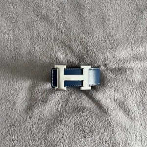 Hermès Bälte  - Säljer ett vändbart Hermès-bälte i blått och svart skinn med en ikonisk silverfärgad H-spänne. Bältet har vit kontrastsöm längs kanterna och är tillverkat i exklusivt läder. Perfekt accessoar för att lyfta din outfit med en lyxig touch.