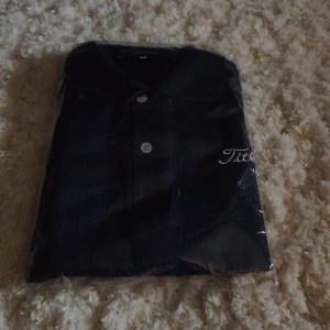 Mörkblå skjorta från Titleist - Helt ny snygg mörkblå skjorta från Titleist med diskreta ränder och klassisk krage. Skjortan har knappar framtill och broderad logga på bröstet och ärmen. Perfekt för dig som gillar sportig och stilren look.