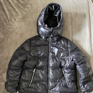 Svart pufferjacka från Lager 157 XS - Snygg svart pufferjacka från Lager 157 i glansigt material. Jackan har huva, två dragkedjeförsedda fickor och hel dragkedja framtill. Perfekt för kalla dagar och ger en cool vibe till din outfit.