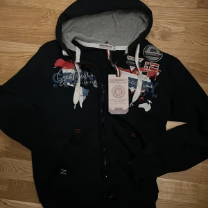 Mörkblå hoodie från GeoNorway Expedition - Mörkblå från GeoNorway Expedition med tryck och broderade märken på bröstet. Tröjan har dragkedja, huva med vit snörning och grått innerfoder. Färgglada detaljer i rött, vitt och blått samt världskarta-tryck framtill. Perfekt för dig som gillar streetwear.