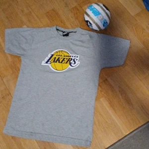 Grå Nike Lakers t-shirt - Snygg grå t-shirt från Nike med stort Los Angeles Lakers-tryck på bröstet. Klassisk rund hals och korta ärmar. Perfekt för dig som älskar basket och vill ha en sportig stil. Materialet är mjuk bomull som känns skönt mot huden.