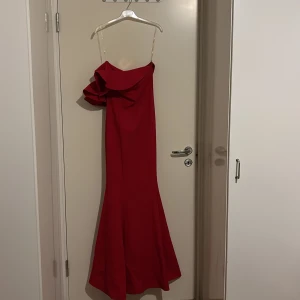 Röd långklänning från La Sarte - Säljer en röd långklänning från La Sarte med asymmetrisk axel och stor volangdetalj framtill. Klänningen har smala axelband och en elegant, figurnära passform som faller snyggt mot golvet. Perfekt för dig som vill sticka ut med en dramatisk look.