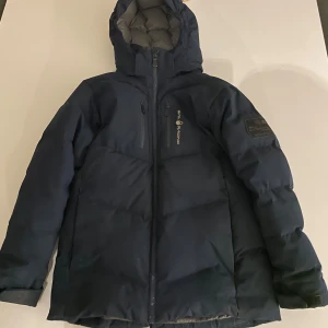 Mörkblå vinterjacka Sail Racing - Snygg mörkblå vinterjacka från Sail Racing med huva och flera dragkedjefickor. Jackan har en clean look med logga på bröstet och patch på ärmen. Perfekt för kalla dagar och riktigt skön att bära. Har en liten skada längs armen(syns på bild 5) samma på båda armarna men inget jätte syndlig.