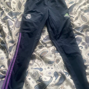 Adidas Real Madrid träningsbyxor  - Mörkblå träningsbyxor från Adidas med Real Madrid-logga och grönt Adidas-märke framtill. Lila ränder längs sidorna och elastisk midja med snörning. Byxorna har smal passform och är gjorda i polyester, perfekta för fotbollsträning. Passar även som S
