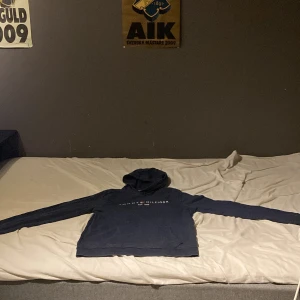 Mörkblå hoodie från Tommy Hilfiger - Säljer en mörkblå hoodie från Tommy Hilfiger med klassisk logga och text på bröstet. Tröjan har huva och långa ärmar, perfekt för dig som gillar en stilren och enkel look. Passar dig som vill ha något bekvämt och snyggt till vardags.