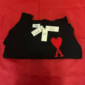 Svart stickad tröja från Ami Paris - Svart stickad tröja från Ami Paris med rund halsringning och ribbade muddar. På bröstet finns ett rött hjärta med bokstaven A, vilket ger en unik och trendig look. Perfekt för dig som vill ha något stilrent men ändå sticker ut.