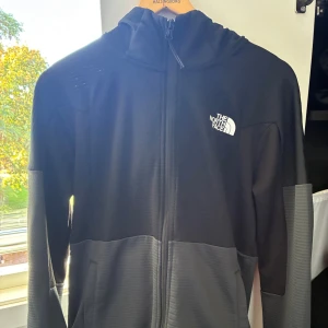 Svart och grå kofta från The North Face - Snygg svart och grå kofta med zip från The North Face med huva och hel dragkedja. Koftan har två sidofickor och logga på både bröstet och ryggen. Perfekt för dig som gillar en sportig och clean look.