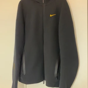Nike nochta - Svart hoodie från Nike med gul Swoosh på bröstet och små gula broderade detaljer på ryggen. Tröjan har huva, långa ärmar och två fickor med dragkedja framtill. Perfekt för dig som gillar sportig och clean stil.