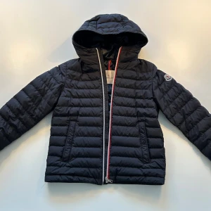 Svart jacka med huva från Moncler - Svart Moncler-jacka i storlek 12A/152cm. Mycket fint skick, inga tecken på slitage.