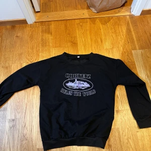 Svart Corteiz sweatshirt med tryck - Svart sweatshirt från Corteiz med stort vitt tryck på bröstet där det står 'Corteiz Rules The World' och en grafisk illustration. Tröjan har rund hals, långa ärmar och ribbade muddar vid ärmslut och nederkant. Perfekt för dig som gillar streetwear och snygga prints.