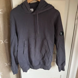 Storlek M men mer som en S. Mörkgrå hoodie från C.P. Company. - Snygg mörkgrå hoodie från C.P. Company med klassisk logga på ärmen och tryck på luvan. Tröjan har huva med snörning, stor magficka och ribbade muddar. Perfekt för dig som gillar streetwear och vill ha en stilren look.