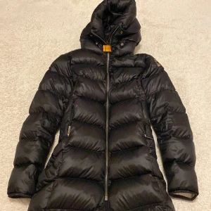 Svart lång dunjacka Parajumpers - Säljer en svart lång dunjacka från Parajumpers. Jackan har en hög krage, huva, dragkedja framtill och den klassiska gula detaljen vid halsen. Tjockt vadderad för extra värme och med logga på ärmen. Perfekt för kalla dagar. Knappt använt, köpt fel storlek. 