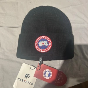 Svart ribbad mössa från Canada Goose - Svart ribbstickad mössa från Canada Goose med klassisk rund logopatch framtill i rött, vitt och blått. Mössan har uppvikt kant och är tillverkad i mjukt material som håller dig varm. Perfekt för kalla dagar och snygg till streetwear. ÄKTA KÖPT FRÅN FARFETCH aldrig använd 
