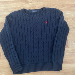 Mörkblå stickad tröja Polo Ralph Lauren - Snygg mörkblå stickad tröja från Polo Ralph Lauren i storlek XS. Tröjan har kabelstickat mönster och en röd broderad logga på bröstet. Rund halsringning och långa ärmar med ribbade muddar. Perfekt för dig som gillar klassisk och stilren look.