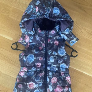 Blommig dunväst från H&M med huva - Snygg dunväst från H&M med blommigt mönster i blått, rosa och lila toner. Västen har en avtagbar huva med tryckknappar och dragkedja framtill. Perfekt för att ge outfiten lite extra färg och stil under kyliga dagar.