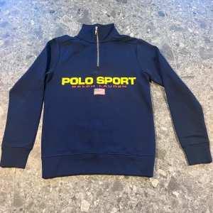 Marinblå Polo Sport half zip tröja - Snygg marinblå tröja från Polo Sport Ralph Lauren med gul och röd logga framtill och amerikansk flagga. Half zip-dragkedja i halsen och långa ärmar.