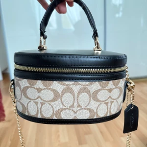 Coach oval handväska med kedja - Snygg oval handväska från Coach med beige och brunt mönster, svarta skinn-detaljer och guldfärgad kedja. Väskan har dragkedja runt om och ett klassiskt Coach-tag. Perfekt för dig som vill ha något unikt och trendigt.