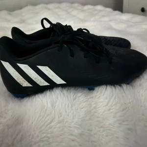 Adidas Predator svarta fotbollsskor - Svarta Adidas Predator fotbollsskor med klassiska vita ränder på sidan och texten 'Predator' på hälen. Skorna har snörning och är tillverkade i syntetmaterial med låg profil och platt sula för bra grepp på planen.
