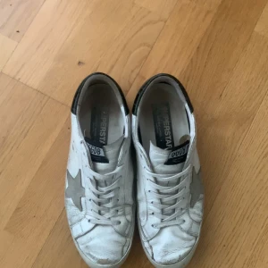 Golden Goose vita sneakers med stjärna - Golden Goose sneakers i vitt skinn med grå mockadetalj i form av en stjärna på sidan. Skorna har svarta detaljer vid hälen, vita snören och svart sula undertill. Klassisk låg modell med rund tå och platt sula. Perfekta för en cool och avslappnad stil.