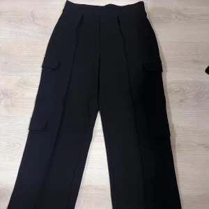 Svarta cargobyxor - Svarta cargobyxor med hög midja och raka ben. Byxorna har stora fickor på sidorna och markerade pressveck framtill.