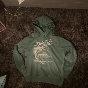 Grön hoodie Polo Ralph Lauren - Säljer en grön hoodie från Polo Ralph Lauren med tryck på bröstet där det står 'Polo R.L. Key West' och en stor fisk och båt. Tröjan har aldrig använts. Perfekt för dig som gillar stockhomstill/grisch och vill ha något unikt.