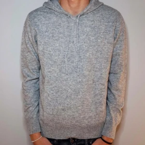 Grå stickad hoodie med huva - Mysig grå stickad hoodie med huva och snörning. Tröjan har lång ärm och ribbade muddar vid ärmslut och nederkant. Perfekt för kyliga dagar och enkel att matcha med jeans eller joggers.