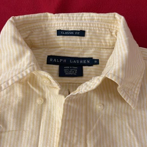 Gulrandig skjorta från Ralph Lauren - Snygg klassisk skjorta från Ralph Lauren i ljusgul och vit randigt mönster. Skjortan har lång ärm, knappar hela vägen och ikoniska broderade loggan på bröstet. Tillverkad i mjuk bomull och har en klassisk fit med krage.