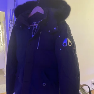 Svart dunjacka Moose Knuckles S - Säljer en svart dunjacka från Moose Knuckles med fet pälskantad huva och ikonisk logga på ärmen. Jackan har dragkedja, knappar och flera fickor. Perfekt för kalla dagar och riktigt snygg streetstil.