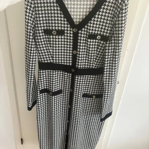 Svartvit långklänning med knappar - Snygg långklänning i svartvitt hundtandsmönster med markerad midja, dekorativa knappar framtill och fyra fickdetaljer. Klänningen har lång ärm och v-ringning med svarta kanter som ger en klassisk och trendig look.
