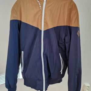 Blå och beige vindjacka med huva - Snygg tvåfärgad vindjacka i marinblått och beige med vit dragkedja och vita detaljer vid fickorna. Jackan har huva, ribbade muddar och en sportig look. Perfekt för dig som gillar stilren och avslappnad streetwear.