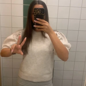 Beige stickad tröja med puffärm - Säljer en beige stickad tröja med korta puffärmar och rund hals. Tröjan har en mjuk och mysig känsla och ribbade kanter nedtill och vid ärmslut. Perfekt för dig som gillar en enkel men trendig look.