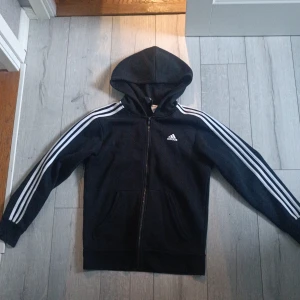Svart Adidas hoodie med dragkedja - Svart hoodie från Adidas med klassiska vita ränder längs ärmarna och Adidas-logga på bröstet. Tröjan har huva, hel dragkedja och två fickor framtill. Perfekt för en sportig look och chill vardagsstil.