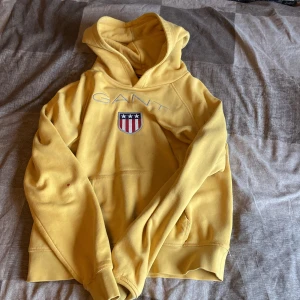 Gul hoodie från GANT med broderad logga - Gul hoodie från GANT med broderad logga och sköld på bröstet. Tröjan har huva, känguruficka och långa ärmar. Mjuk bomullskvalitet och klassisk passform, perfekt för en avslappnad stil.