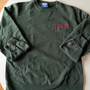 Mörkgrön sweatshirt från Champion - Mörkgrön sweatshirt från Champion i storlek medium. Tröjan har broderad Champion-logga i rött och grått på bröstet samt en liten logga på ärmen. Rund hals och ribbade muddar vid ärmslut och nederkant. Köpt av mig i Tokyo! Är öppen för prisförslag