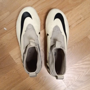 Nike beige fotbollsskor - Säljer ett par beiga fotbollsskor från Nike med stor svart Nike logga på ovansidan och coolt svart mönster på sidan. Skornahar platt sula med dobbar och är tillverkade i syntetmaterial. De har en smidig design utan snörning och en praktisk flik vid hälen.
