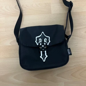 Svart axelväska från Trapstar - Säljer en svart axelväska från Trapstar med unik vit och svart spänndetalj på locket. Väskan har en justerbar rem och textur i materialet som ger en cool vibe. Märket syns tydligt på både rem och etikett. Perfekt för att bära dina essentials.