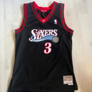 Svart Sixers baskettröja Iverson #3 - Säljer en svart Philadelphia 76ers baskettröja med Iverson och nummer 3 på ryggen. Tröjan är ärmlös med röd och vit kant, mesh-material och klassisk NBA-logga på bröstet. Perfekt för dig som gillar retro och sport. Köpt från basketshoppen och är helt oanvänd. Den är unisex och storlek xs. 