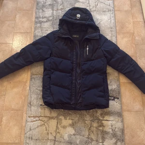 Mörkblå pufferjacka från Sail Racing  - Snygg mörkblå pufferjacka från Sail Racing med huva och dragkedja framtill. Jackan har två bröstfickor med dragkedja och logga på bröstet samt på huvan. Perfekt för kalla dagar och har en modern, sportig look. 